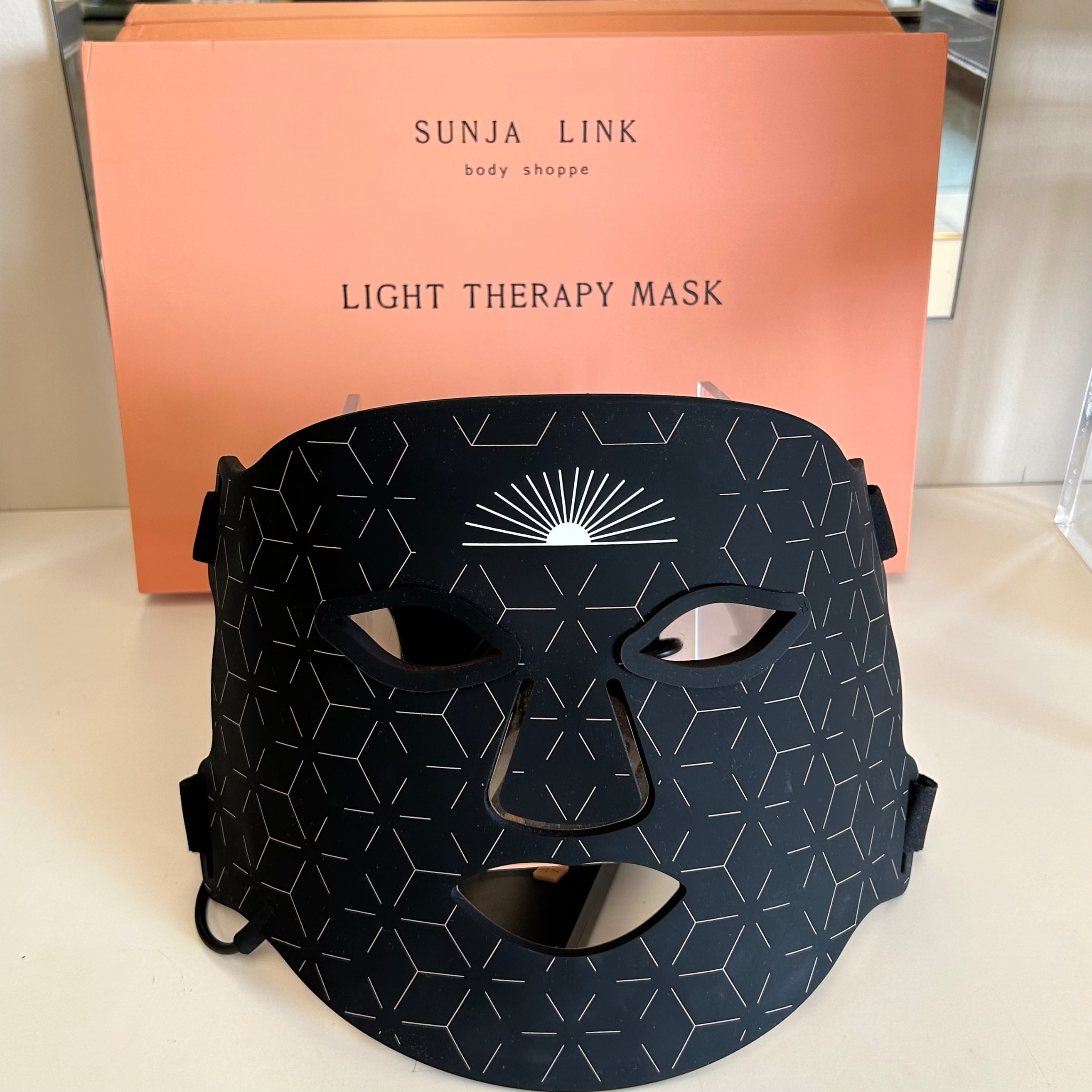 A black light therapy face mask with white geometric patterns and a sunburst design, displayed in front of its peach-colored Sunja Link packaging box.