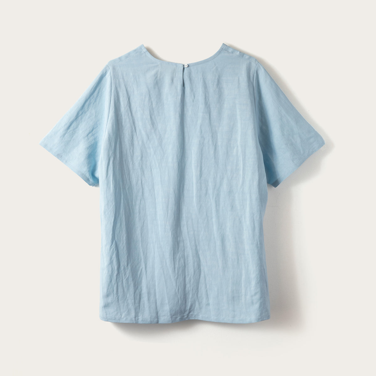 A light blue short-sleeve top with a relaxed, slightly wrinkled texture, shown from the back against a warm off-white background.