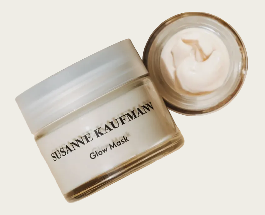 A jar of Susanne Kaufman Glow Mask with its lid off, showing the creamy off-white mask inside, set on a light wood background.