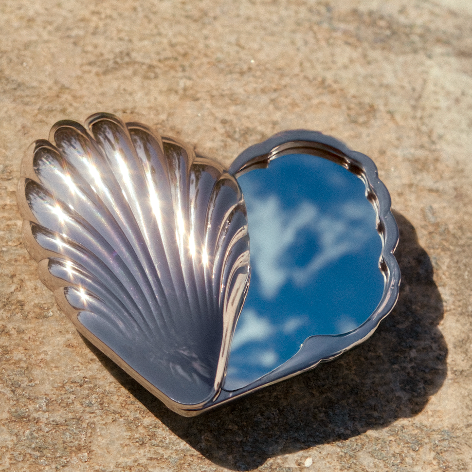 A polished silver-toned metal tray shaped like a clam shell, photographed outdoors on a sandy surface. The reflective interior shows a blue sky with white clouds.