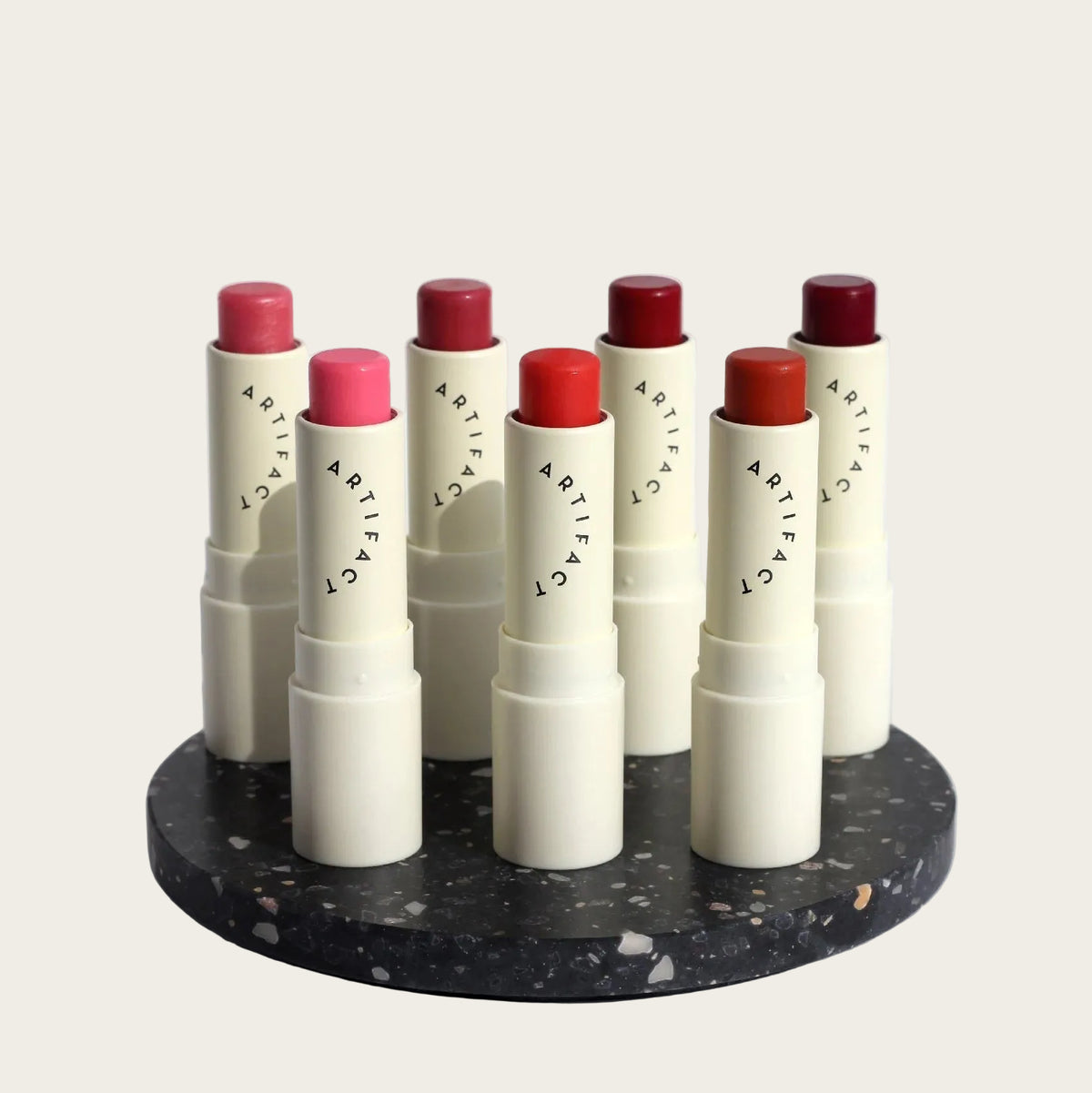 Six lipstick tubes from the brand ARTIFACT arranged on a round black-and-white speckled tray against a clean white background. The lipsticks come in pink, coral, red, and burgundy shades, with the cream-colored tubes showing the ARTIFACT logo.