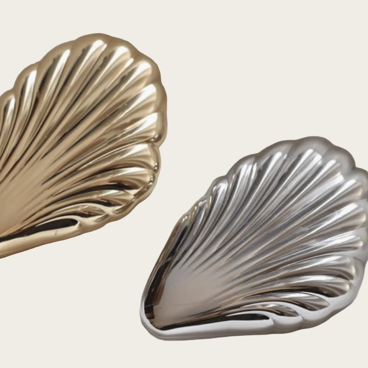 Two metallic shell-shaped accessories, one in polished gold and one in polished silver, displayed on a light beige background.