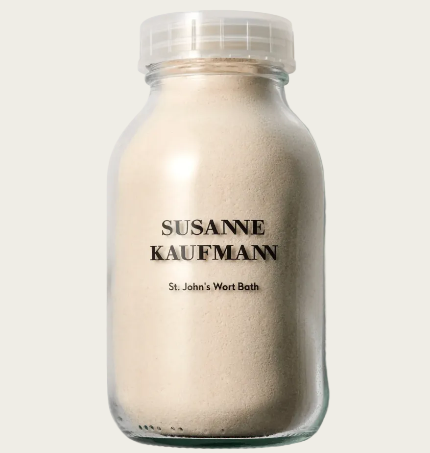 A clear glass bottle filled with a light beige powder, featuring a clear ribbed cap and black text that reads 'SUSANNE KAUFMANN' and 'St. John's Wort Bath'.