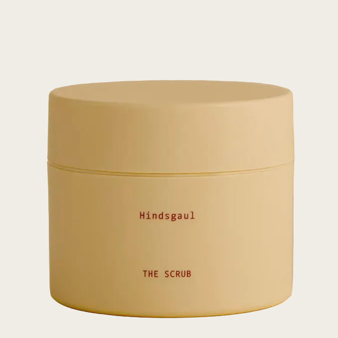 A cream-colored cosmetic jar with a matching lid, featuring the brand name 'Hindsgaul' and product name 'THE SCRUB' printed in a dark red typewriter font, set against a matching cream background.